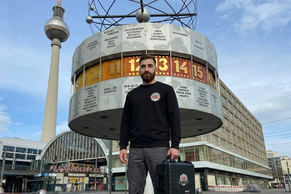 Ein Servicetechniker von MK steht mit einem Werkzeugkoffer vor der Weltzeituhr auf dem Alexanderplatz in Berlin, im Hintergrund ist der Fernsehturm zu sehen.