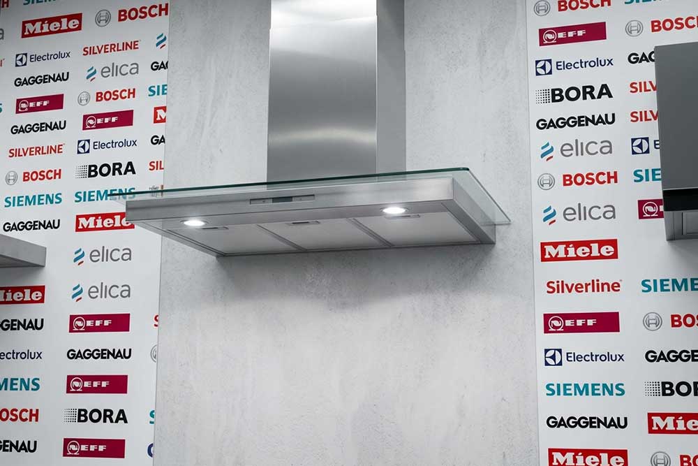Eine Dunstabzugshaube aus Edelstahl und Glas ist an einer Wand montiert, die mit Logos von Herstellern wie Miele, Bosch, Bora, Gaggenau, Siemens und Elica bedruckt ist.