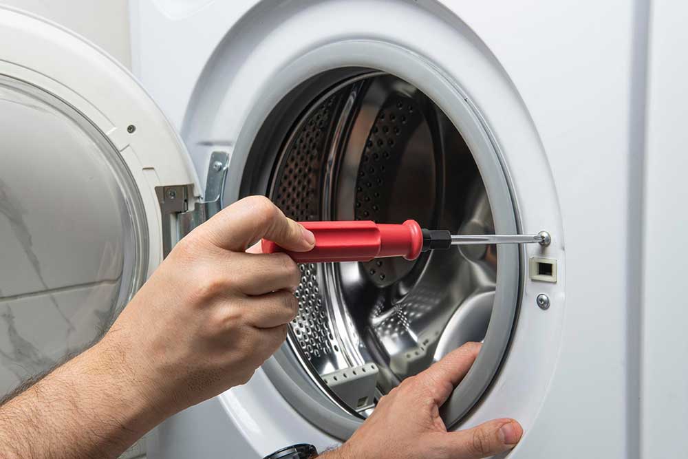 Detailaufnahme einer Hand, die mit einem roten Schraubendreher den Schließmechanismus an der Tür eines weißen Wäschetrockners repariert.