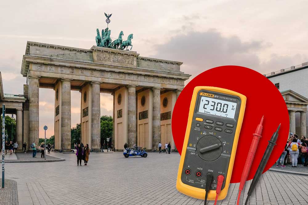 Ein gelbes digitales Multimeter, das eine Spannung von 230,0 Volt anzeigt, ist vor der Kulisse des Brandenburger Tors in Berlin platziert.